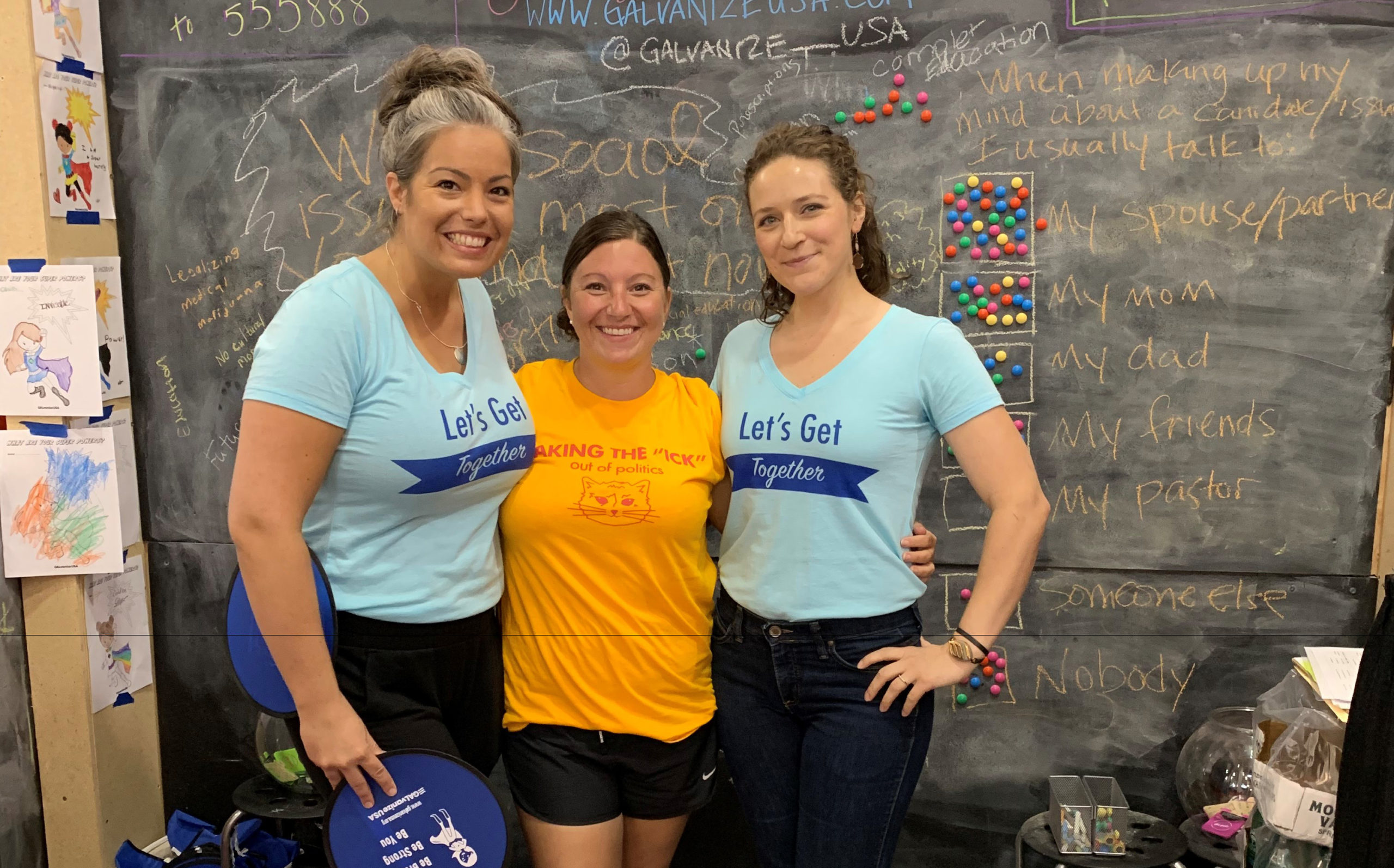 Photo of 3 women smiling with arms around one another standing in front of a chalk board with writing and colored magnets on it.