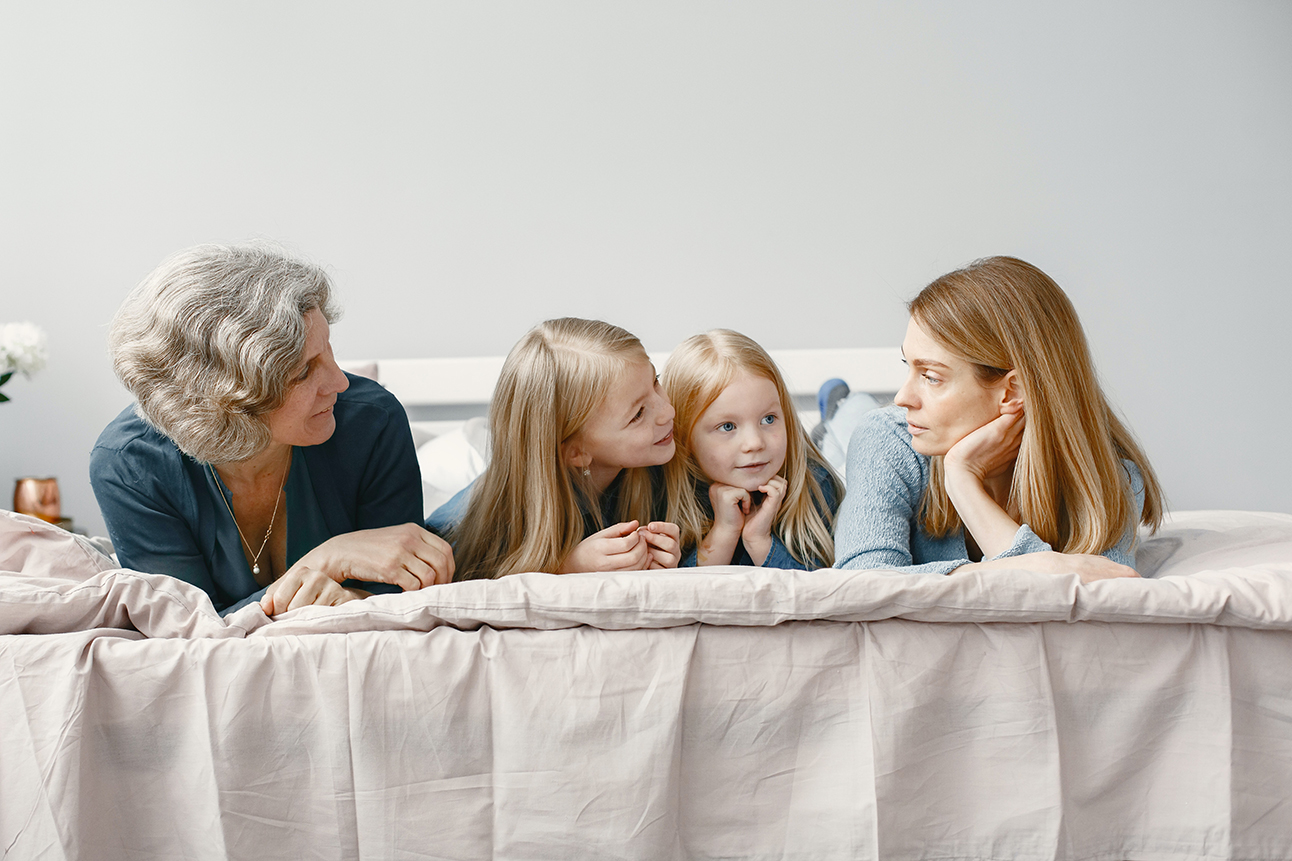 A mom, grandma, and two young girls relax together on a bed